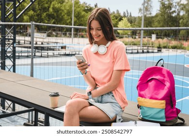 Pretty high school girl teenager pupil student in casual wear with bag sitting in sport yard basketball court holding smart phone using mobile app play game posting chatting texting browsing internet - Powered by Shutterstock