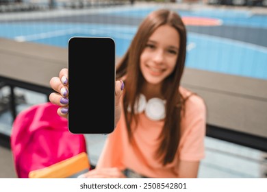 Pretty high school girl teenager pupil student in casual wear with bag headphone sitting in sport yard basketball court using holding smart phone showing mock up blank screen looking at camera - Powered by Shutterstock