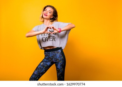 Pretty Hipster Woman Making Heart By Her Hands, Posing At Studio Yellow Background, Wearing Yoga Pants And Funny T-shirt.