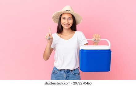 una mujer hispana guapa sonriendo y luciendo amigable, mostrando el número uno y sosteniendo un refrigerador portátil