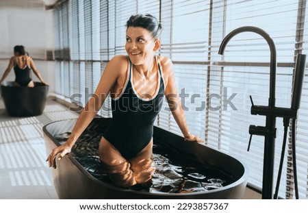 Pretty hispanic young woman in swimsuit deeming in bath with cold water looks aside toothy smiles at bathroom. TannedAmerican girl enjoying vacation at resort at hotel room. Leisure, travel.