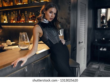 Pretty Hot Woman In Fashion Black Party Dress Leaning On The Bar Counter, Tender Hand Lying On The Waist. Warm Yellow Light Falling From The Back. Stylish Makeup And Curly Hair. Slim Body.