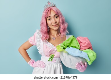 Pretty Housemaid With Pink Dyed Hair Collects Dirty Laundry In Plastic Basin Wears Crown White Dress Gloves Busy Doing Daily Domestic Work Poses Against Blue Background. Professional Cleaning