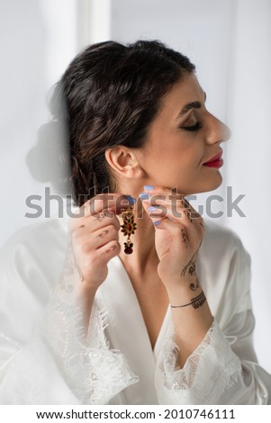 pretty indian bride wearing earring while getting ready for wedding on white