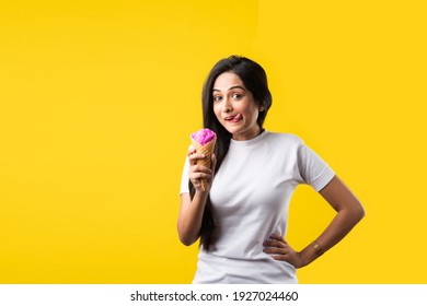 Pretty Indian girl or young Asian woman eating strawberry ice cream in cone against yellow studio background