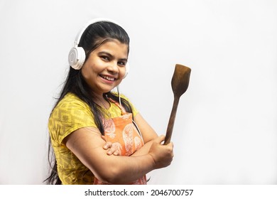 A Pretty Indian Housewife Woman Wearing Cooking Apron And Headphone With Wooden Spatula In Hand Smiles On White Background