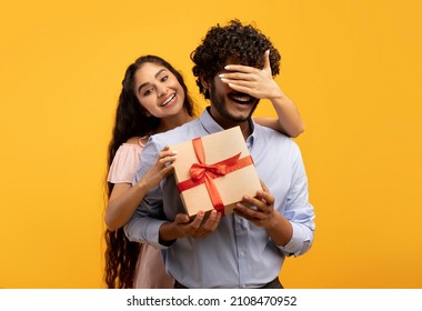 Pretty indian woman covering her boyfriend's eyes, holding wrapped gift box and greeting him with birthday or anniversary, standing over yellow studio background - Shutterstock ID 2108470952
