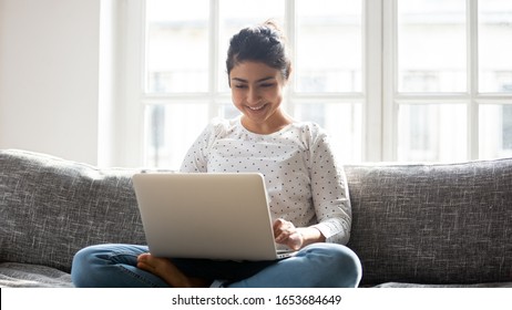 Pretty indian woman settled comfortably on sofa in warm living room holding on lap laptop typing email do remote job, chatting on-line with friend, shopping purchasing enjoy internet sale shop concept Foto Stock