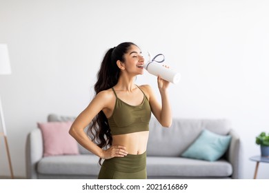 Pretty Indian woman in sports clothes drinking water or protein shake from bottle at home. Millennial Asian lady losing weight, keeping hydrated after workout, taking care of her body indoors - Shutterstock ID 2021616869