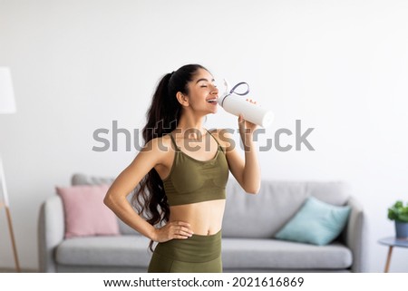 Pretty Indian woman in sports clothes drinking water or protein shake from bottle at home. Millennial Asian lady losing weight, keeping hydrated after workout, taking care of her body indoors