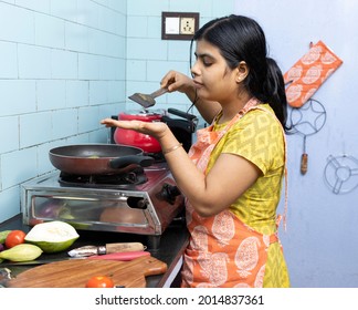 A Pretty Indian Young Woman Wearing Apron Cooking And Tasting Food In Domestic Kitchen On Gas Stove