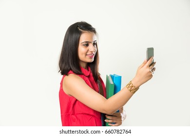 Pretty Indian/Asian College Girl Holding Books And Clicking Selfie With Smartphone, Standing Isolated Over White Background