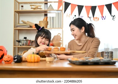 Pretty And Joyful Little Asian Girl In Fancy Costume Making Halloween Cupcakes For The Halloween Party With Her Mom.