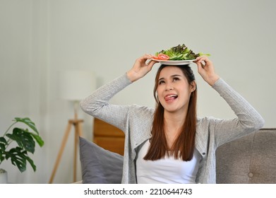 Pretty And Joyful Young Asian Female Putting A Plate Of Her Green Salad On Her Head, Making A Yummy Face, Sitting In Her Living Room.