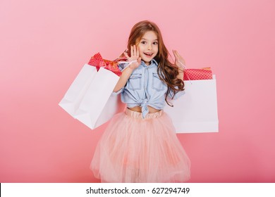 Pretty joyful young girl in tulle skirt, with long brunette hair walking with white packages on pink background. Lovely sweet moments of little princess, pretty friendly child having fun to camera