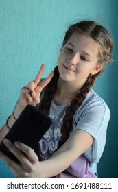 Pretty kid girl 11-12 years old in blue T-shot hold in hand cell phone, taking selfies and showing peace gesture, isolated on turquoise background. Emotional portrait