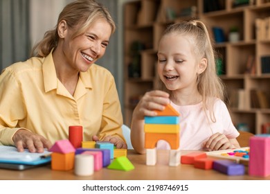 Pretty Kid Little Girl And Female Child Development Specialist Woman Playing With Colorful Wooden Bricks, Sitting At Table And Making Pyramid Together, Smiling