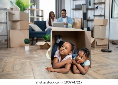 Pretty Kids Lying Inside Cardboard Box While Parents Sitting On Couch And Using Laptop. African Family Enjoying Moving To New Apartment. Concept Of Relocation.