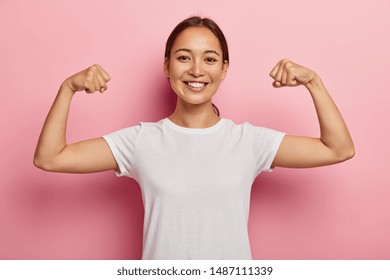 Pretty korean female model keeps fit and healthy, raises hands and shows muscles, feels proud about her achievements in gym, smiles broadly, dressed in white casual wear, poses indoor shows real power