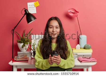 Pretty korean female scholar in green sweater, holds takeawy coffee cup, prepares research at home, poses against cozy workplace with flowers, desk lamp, textbooks and folders. Pink background