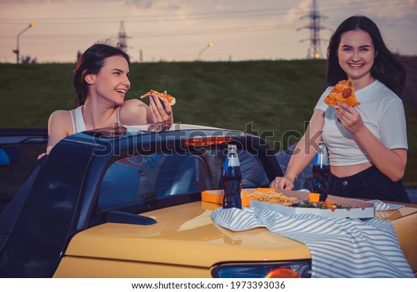 Pretty ladies smiling, eating pizza, posing in\
yellow car cabrio with french fries and soda in glass bottles on\
its trunk. Fast food. Copy\
space