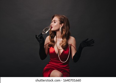 Pretty Lady In Festive Gorgeous Outfit Drinks White Wine. Superstar Woman In Luxury Red Dress Holding Champagne Glass On Black Background