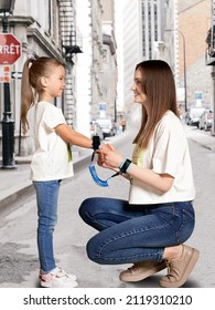 A Pretty Lady In Jeans And A T-shirt Is Sitting Down With A Standing Girl On The Street. The Woman And The Child Are Connected By A Blue Safety Anti Lost Leash With Soft Wristbands And Key Lock.