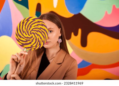 Pretty lady posing covering eyes round giant lollipop at colored wall, pensive looking at camera. Lovely woman in beige suit with candy on stick. Summer fashion concept. Copy ad text for space poster