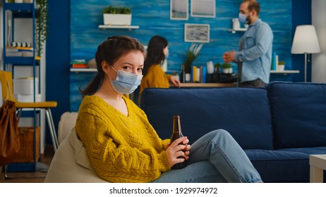 Pretty Lady Taking Off Protection Face Mask Looking At Camera Drinking Beer Spending Good Time With Friends In Living Room Respecting Social Distance. People Socializing During Covid 19 Outbreak