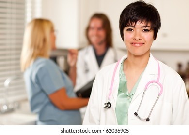 Pretty Latino Doctor Smiles At The Camera As Colleagues Talk Behind Her.