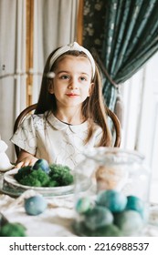 Pretty Little Armenian Girl Helps With Baking For Easter On Veranda On Sunny Spring Day Decorated With Flowers And Easter Decor, Eggs, Cake And Willow Branches, Easter Family Celebration