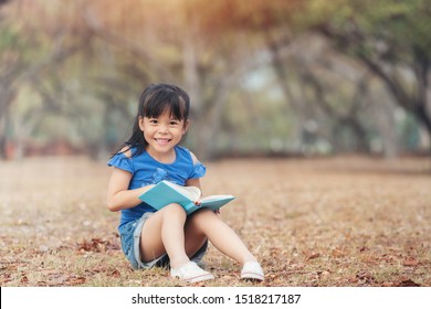 Pretty Little Asia Children Girl Black Hair In Blue Shirt Sitting Reading A Book On Brown Grass Field. Children And Science Education Concept.