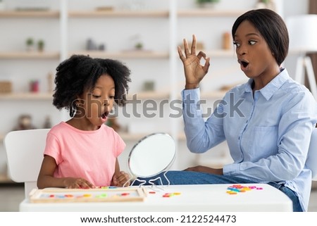 Pretty little black girl attending speech therapy session at clinic, looking at mirror, beautiful young african american woman speech-language pathologist working on sounds with kid, articulating