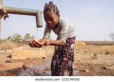 Pretty little black girl hunched over by a West African village pump, happily holding both hands to catch the fresh water running out of the pipe; water scarcity concept