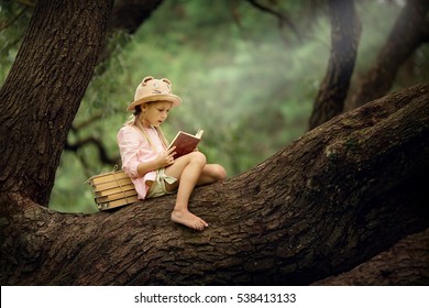 A pretty little blonde girl in a straw hat reading a book on a large spreading tree. Children and science.