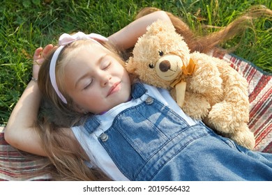 Pretty Little Child Girl With Closed Eyes Laying Down With Her Teddy Bear Toy On Blanket On Green Grass In Summer Taking A Nap.