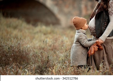 pretty little girl in brown hat and coat standing in the meadow in the park and kiss stomach of her pregnant mother at autumn 