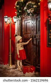 Pretty Little Girl Is Celebrating Christmas And Giving Red Gift Box. Child On Decorated Street Knocking On Wooden Door In Winter. Fantastic Vintage Christmas Picture