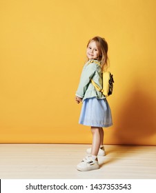 Pretty Little Girl Child In Stylish Dress And Jeans With A Yellow Backpack Behind Her Back Jumping Smiling Looking At The Camera. On Yellow Studio Background