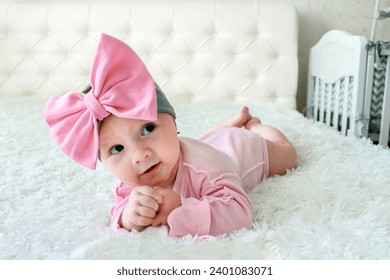 A pretty little girl of European appearance with a huge bow on her head lies on the bed and smiles at the frame. Close-up portrait of a small baby girl, smiling baby at home on the bed - Powered by Shutterstock