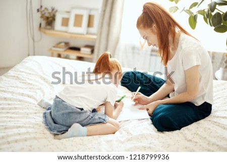 Pretty little girl with ginger ponytail sitting on bed with her mother and using coloured pencils to make Christmas card, interior of cozy living room on background
