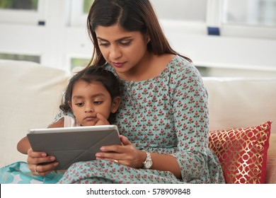 Pretty little girl and her middle-aged mother watching cartoons on digital tablet with interest while sitting in living room
