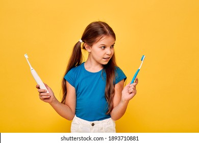 Pretty little girl holds manual and electric sonic toothbrush. Child selects of kind of toothbrush, on yellow background