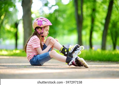 Pretty Little Girl Learning To Roller Skate On Beautiful Summer Day In A Park. Child Wearing Safety Helmet Enjoying Roller Skating Ride Outdoors. Kid Hurt Her Knee While Riding. 