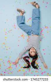 Pretty little girl lying upside down on the blue floor, covered in confetti. She wears have jeans and black and white striped shirt on them.