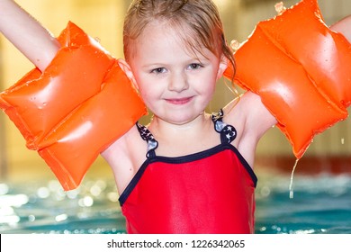 A pretty little girl with orange arm bands smiling and playing happily in the swimming pool, wearing a red swimming costume