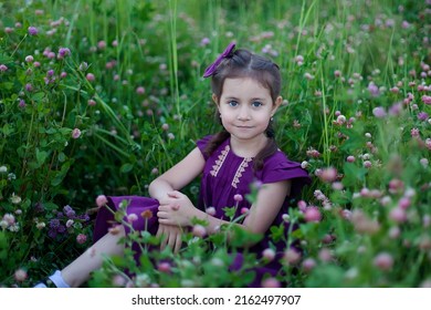 A pretty little girl in a purple dress and a bow on a clover field, picking flowers and enjoying life.