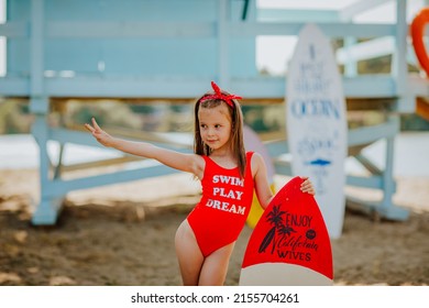 Pretty little girl in red bikini posing with small surfboard like a model on the beach against blue lifeguard tower
