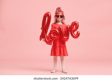 A pretty little girl in a red dress and heart-shaped glasses holds a balloon with love word and smiles joyfully. Portrait on a pink background. Valentine's Day. Mothers Day. Birthday.