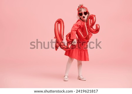 A pretty little girl in a red dress and heart-shaped glasses holds a balloon with love word and smiles joyfully. Portrait on a pink background. Valentine's Day. Mothers Day. Birthday.
